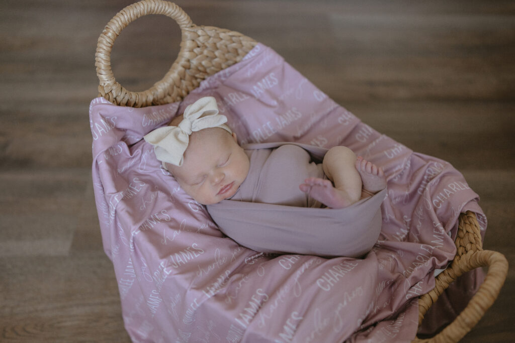 Baby newborn picture in a basket with a personalized name blanket, purple swaddle, and white bow headband